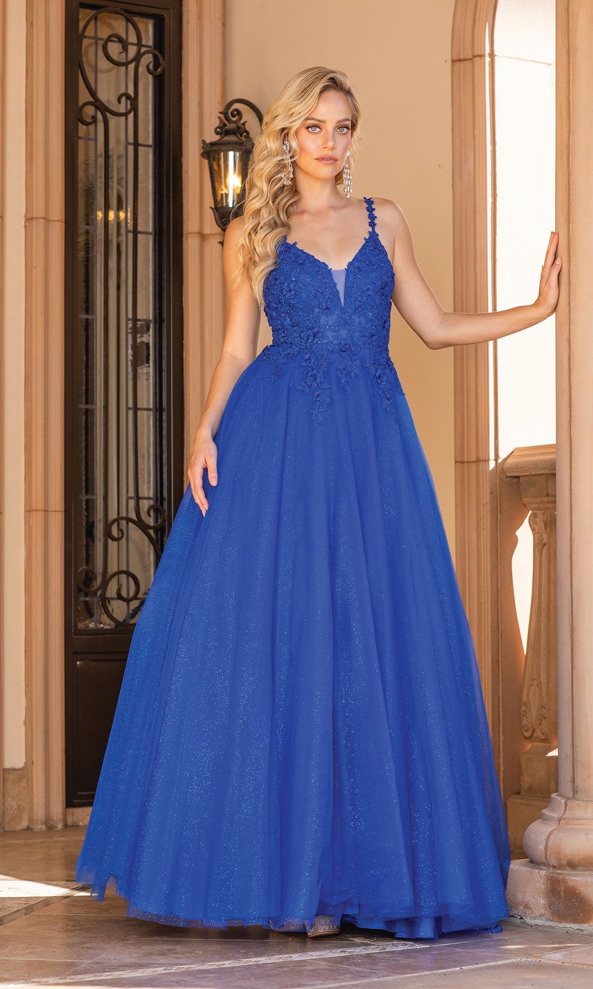 A woman stands in a doorway wearing the Dancing Queen 4328 Long Glitter Prom Dress in royal blue, featuring floral lace details and a fitted bodice—a stunning choice for formal occasions.